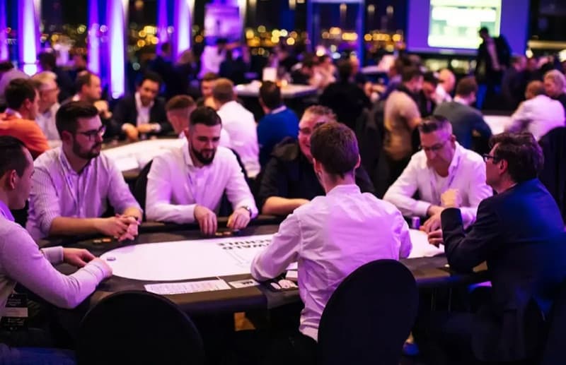 Gäste eines BusinessPokern-Abends sitzen in Hemd und Sakko an mehreren Pokertischen, der Saal ist violett ausgeleuchtet.
