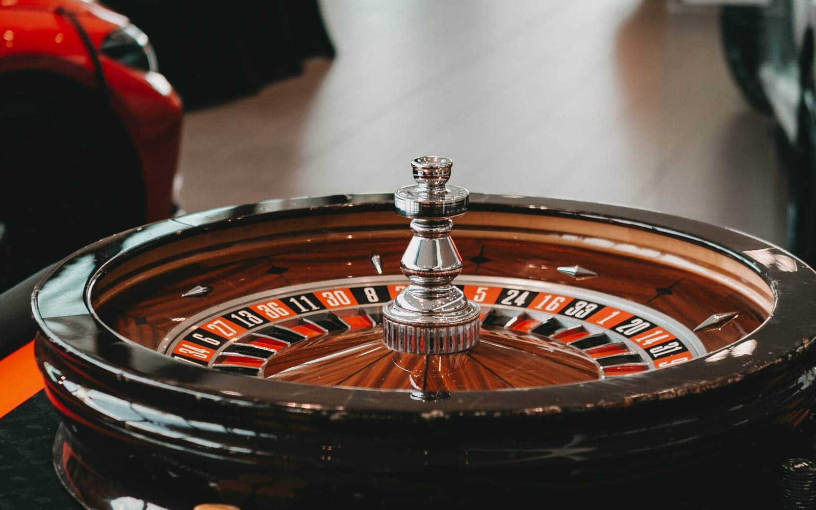 Ein Roulettekessel aus dunklem Holz mit silbernem Drehkreuz auf einem Spieltisch, dahinter unscharf ein roter Sportwagen.
