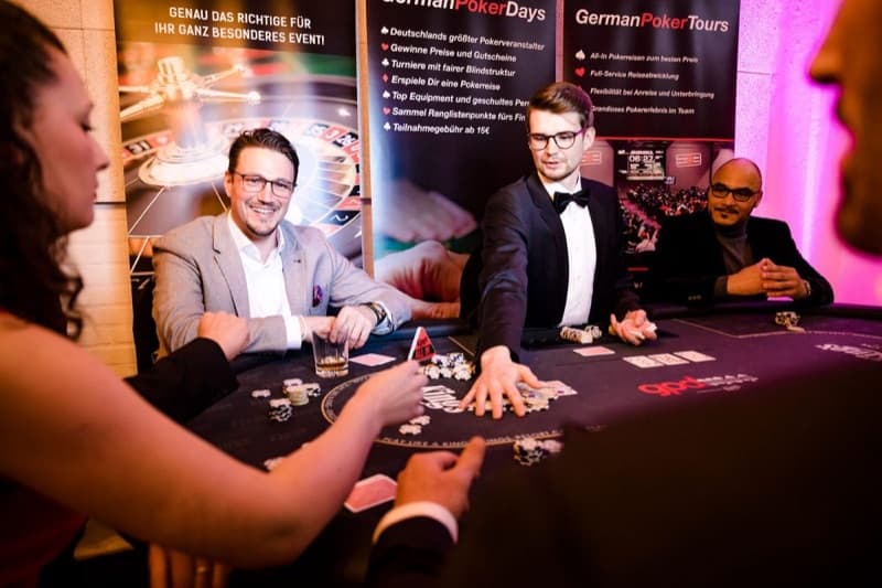 Ein Croupier mit Fliege teilt an einem Pokertisch aus, dahinter stehen Roll-up-Banner der German Poker Days und der German Poker Tours.