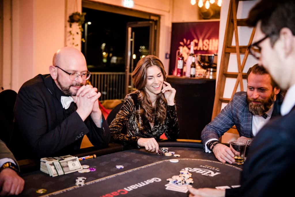 Drei Gäste sitzen an einem Pokertisch und lachen, vor ihnen liegen Chips und Karten.