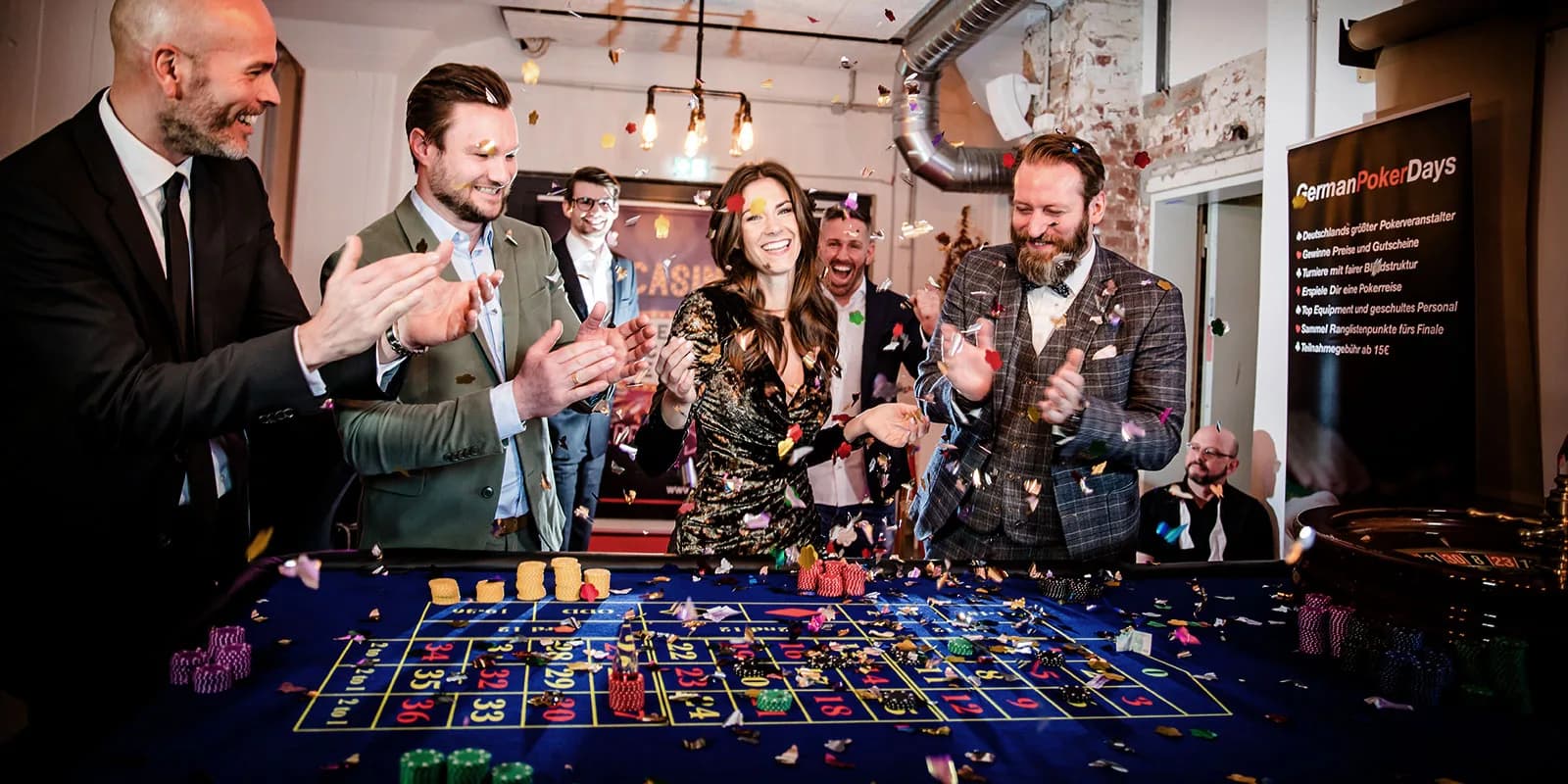 Gäste in Anzug und Abendkleid jubeln im Konfettiregen an einem Roulettetisch, rechts steht ein Banner der German Poker Days.
